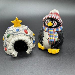 Op de afbeelding: Een keramische set met een iglo en een pinguïnbeeldje. De iglo is wit met kleurrijke lichtjes en een gele ster. De pinguïn is zwart en wit met een rood-witte muts, een geruite sjaal en gele voeten. Voor feestelijke decoratie.