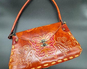 May include: A brown leather handbag with a butterfly and floral design. The bag features a handle, a clasp closure, and decorative stitching along the edges. The butterfly is embossed in the center of the flap.