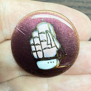 May include: A circular button in maroon with a sailing ship design. The ship is white with gold accents and black outlines. The button is held in a hand.