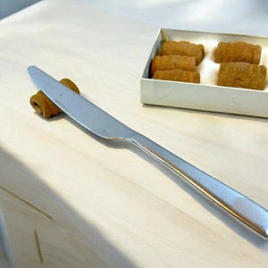 May include: A silver butter knife with a wooden handle is lying on a light wood surface.  Four small, brown, cylindrical pastries are in a white box.