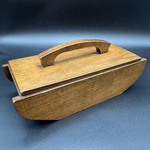 May include: A rectangular wooden box with a medium brown finish and a visible wood grain. The box features a curved bottom base and a flat lid with a single arched handle centered on top. The construction is solid with smooth, clean edges.