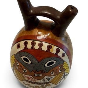 May include: A round ceramic vessel with a dark brown double-spout handle. The body features a painted design with a central gray face, red accents, and yellow geometric patterns on a cream background, including small black bird-like motifs.