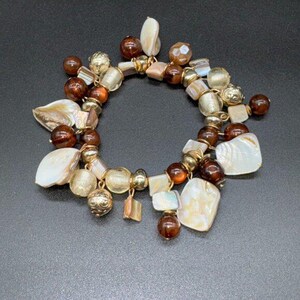 May include: A bracelet with a variety of beads and shell pieces. The bracelet features gold-coloured beads, brown and amber beads, and irregular shell pieces. The bracelet is set against a dark background.