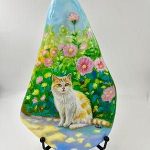 May include: A decorative, hand-painted stone depicting a white and orange cat in a garden with pink flowers. The stone is mounted on a black metal stand. The background shows a blue sky and green foliage.
