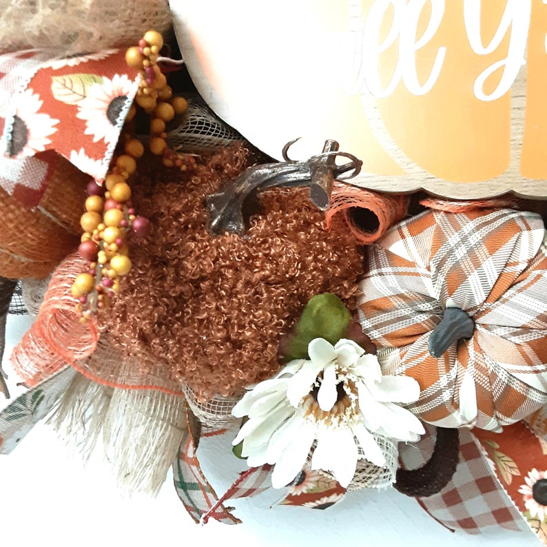 Puede incluir: Una corona con tem&aacute;tica oto&ntilde;al con un gran letrero blanco en forma de calabaza que dice "Happy" en escritura blanca. La corona presenta una calabaza marr&oacute;n texturizada, calabazas a cuadros, girasoles y cintas de color naranja y crema.