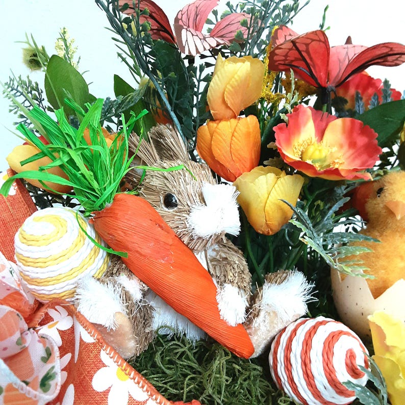May include: An Easter arrangement with a straw rabbit holding a large orange carrot, colourful eggs, and a chick hatching from an eggshell. The arrangement includes orange, yellow, and red flowers, and decorative butterflies.