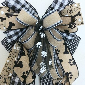 May include: A large, multi-layered bow made from burlap, black and white buffalo check, and black and white paw print ribbon. The bow is tied with a black sheer ribbon with white paw prints.