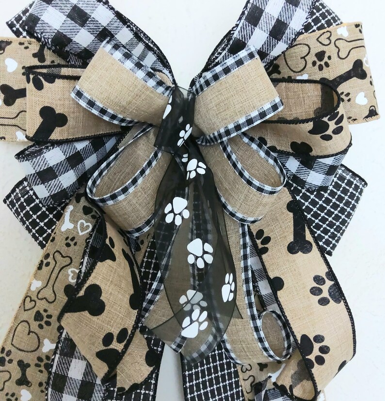 May include: A large, multi-layered bow made from burlap, black and white buffalo check, and black and white paw print ribbon. The bow is tied with a black sheer ribbon with white paw prints.