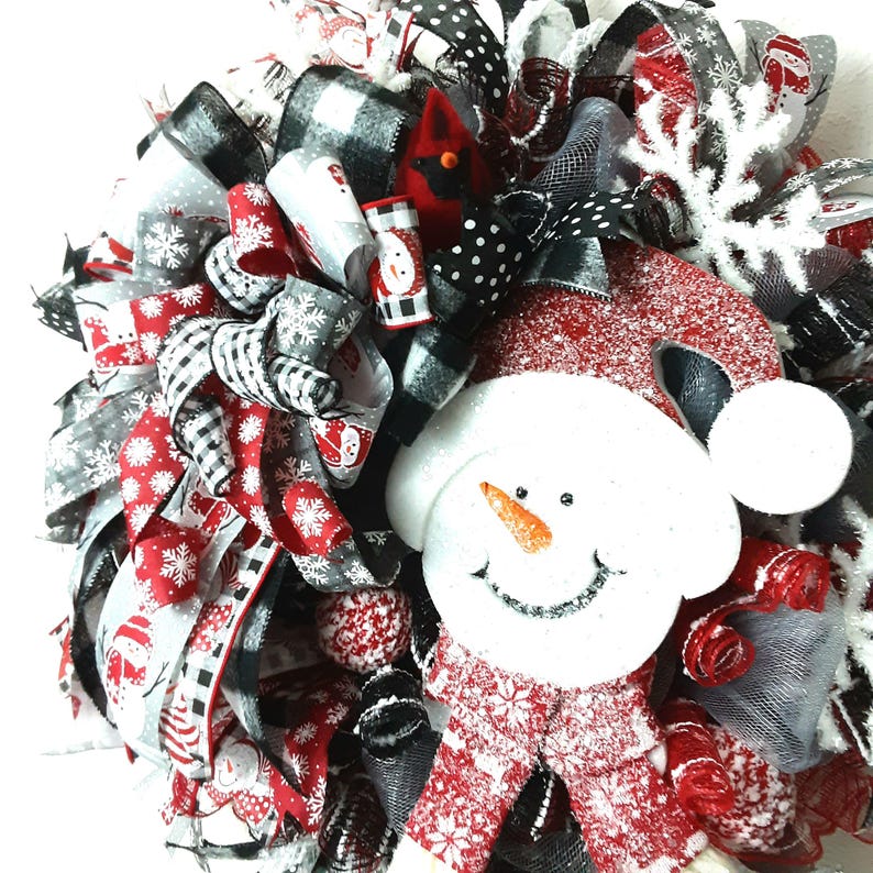Puede incluir: Una corona decorativa con una figura central de mu&ntilde;eco de nieve con cara blanca brillante, nariz de zanahoria naranja y gorro rojo. La corona est&aacute; compuesta por cintas superpuestas en rojo, negro, blanco y gris, con patrones de copos, cuadros y lunares.