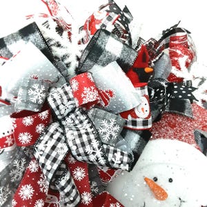Puede incluir: Un lazo decorativo para corona navide&ntilde;a con una mezcla de cintas en rojo, negro, blanco y gris. Los patrones incluyen cuadros escoceses en blanco y negro, copos de nieve blancos sobre fondos rojos y grises, y motivos de mu&ntilde;ecos de nieve.