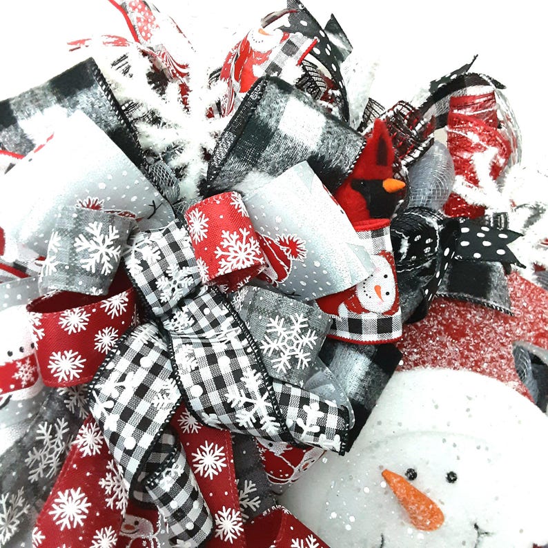 Puede incluir: Un lazo decorativo para corona navide&ntilde;a con una mezcla de cintas en rojo, negro, blanco y gris. Los patrones incluyen cuadros escoceses en blanco y negro, copos de nieve blancos sobre fondos rojos y grises, y motivos de mu&ntilde;ecos de nieve.