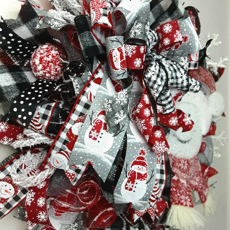 Puede incluir: Una corona decorativa festiva con una mezcla de cintas rojas, negras, blancas y grises. El dise&ntilde;o incluye patrones de cuadros de b&uacute;falo en blanco y negro, lunares blancos y cintas grises estampadas con mu&ntilde;ecos de nieve y copos de nieve.