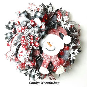Puede incluir: Una corona decorativa con una figura central de mu&ntilde;eco de nieve con gorro y bufanda rojos, rodeada de cintas negras, blancas y rojas con patrones de copos de nieve y cuadros, copos de nieve blancos y el texto CandysWreathShop.
