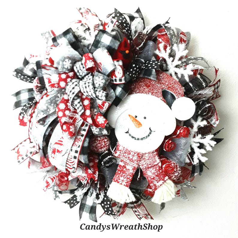 Puede incluir: Una corona decorativa con una figura central de mu&ntilde;eco de nieve con gorro y bufanda rojos, rodeada de cintas negras, blancas y rojas con patrones de copos de nieve y cuadros, copos de nieve blancos y el texto CandysWreathShop.