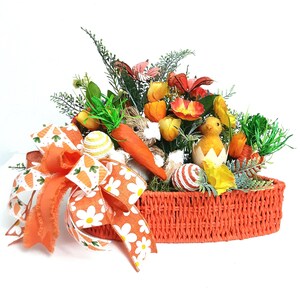 May include: An orange woven basket filled with Easter decorations. The arrangement includes a large orange bow with carrot and daisy patterns, faux flowers, a carrot, a chick hatching from an egg, and decorative eggs.