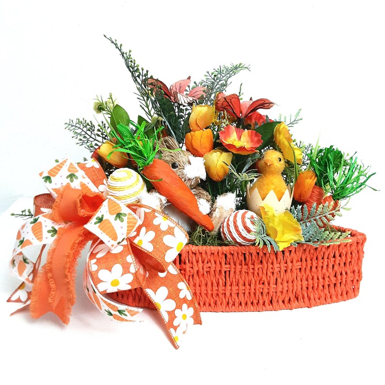 May include: An orange woven basket filled with Easter decorations. The arrangement includes a large orange bow with carrot and daisy patterns, faux flowers, a carrot, a chick hatching from an egg, and decorative eggs.