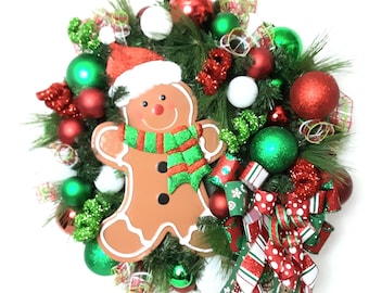 Puede incluir: Una corona navideña festiva con un hombre de jengibre con gorro de Papá Noel y bufanda verde y roja. La corona está adornada con adornos rojos, verdes y blancos, junto con cintas rojas y verdes, creando una alegre exhibición navideña.