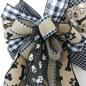 May include: A black and white buffalo plaid ribbon, a black and white paw print ribbon, and a burlap ribbon with black paw prints and bone designs are tied together in a bow. The bow is decorated with a black sheer ribbon with white paw prints.
