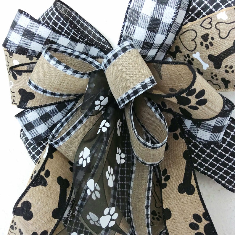May include: A black and white buffalo plaid ribbon, a black and white paw print ribbon, and a burlap ribbon with black paw prints and bone designs are tied together in a bow. The bow is decorated with a black sheer ribbon with white paw prints.