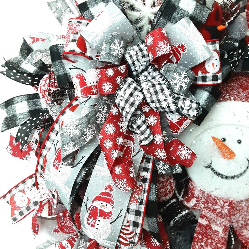 Puede incluir: Una corona decorativa festiva con una cara de mu&ntilde;eco de nieve, nariz de zanahoria y bufanda roja. La corona est&aacute; adornada con m&uacute;ltiples cintas en rojo, blanco, gris y negro, con patrones de mu&ntilde;ecos de nieve, copos y cuadros.