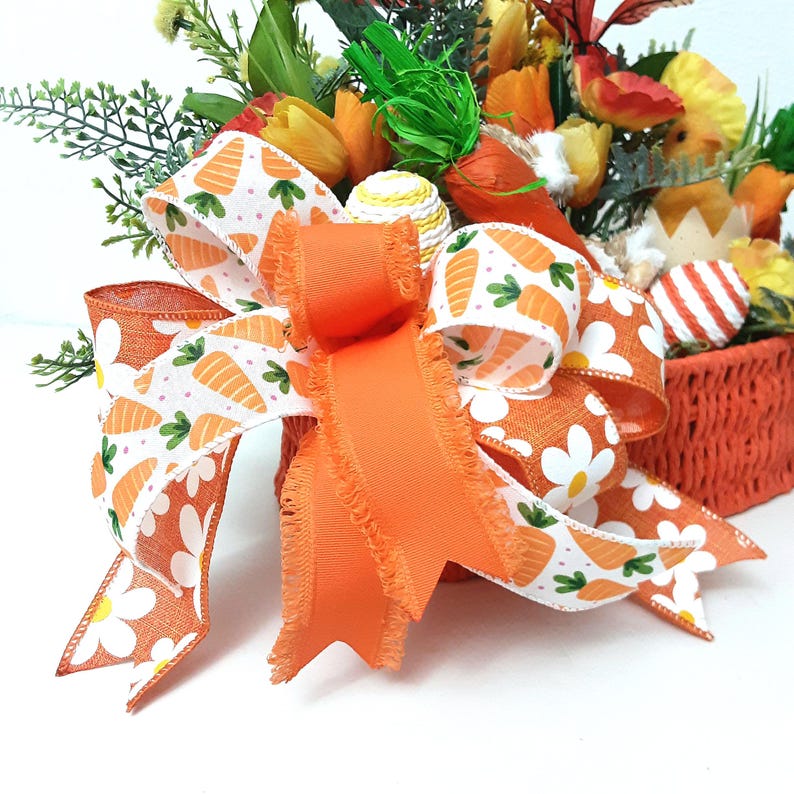 May include: An Easter-themed basket with an orange bow adorned with carrot and daisy motifs. The basket contains colourful flowers, decorative eggs, and artificial carrots, forming a cheerful spring display.