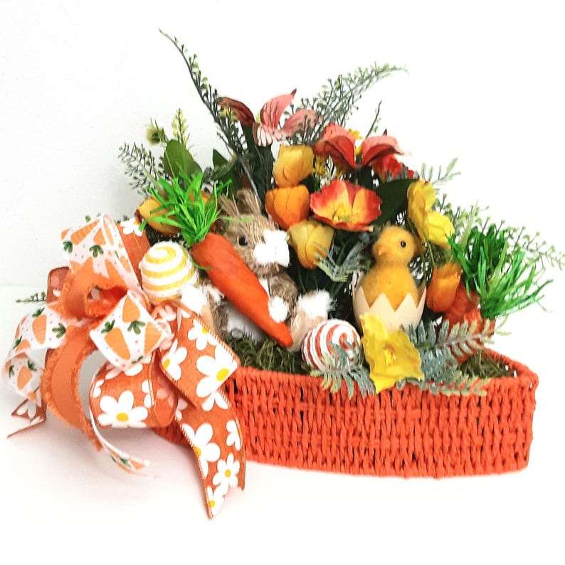 May include: An Easter arrangement in an orange woven basket. It features a plush bunny, a chick hatching from an egg, faux carrots, and colourful flowers. A large orange bow with carrot and daisy patterns adds a festive touch.