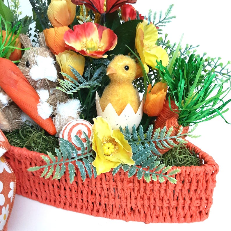 May include: A decorative Easter basket filled with spring-themed ornaments. It includes a fluffy bunny, a yellow chick hatching from an egg, orange carrots, and bright flowers. The basket is woven in a vivid orange, creating a cheerful display.