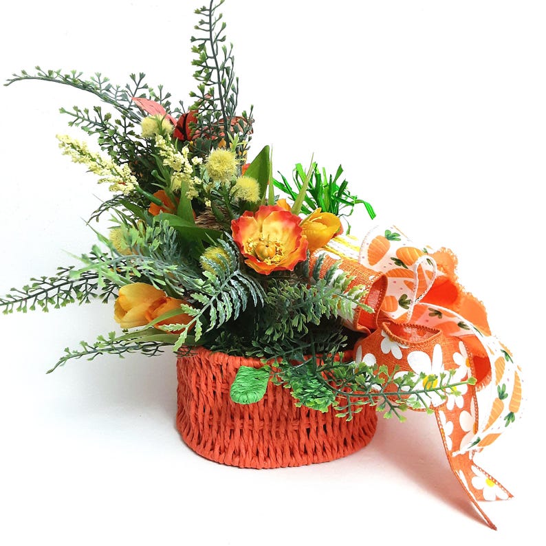 May include: A decorative floral arrangement in an orange woven basket. The arrangement includes orange and yellow flowers, green foliage, and a large orange ribbon with carrot and daisy patterns. The basket is approximately 15 cm in diameter.