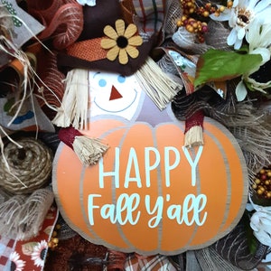 Puede incluir: Una corona de oto&ntilde;o con un dise&ntilde;o de espantap&aacute;jaros y una calabaza con la frase "Happy Fall y'all". La corona incluye paja, arpillera y elementos florales en naranja, marr&oacute;n y blanco.