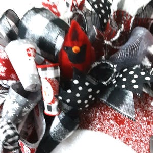 Puede incluir: Una corona decorativa con una figura central de cardenal rojo rodeada de cintas a cuadros blancos y negros, cintas negras con lunares blancos y cintas rojas con motivos de copos de nieve, acentuada con un peque&ntilde;o estampado de mu&ntilde;eco de nieve.