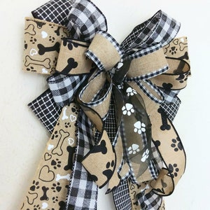 May include: A black and white buffalo plaid ribbon, a black and white paw print and bone ribbon, and a brown burlap ribbon are tied together in a large bow. The bow is decorated with a sheer black ribbon with white paw prints.