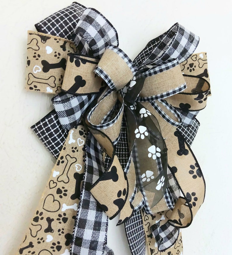 May include: A black and white buffalo plaid ribbon, a black and white paw print and bone ribbon, and a brown burlap ribbon are tied together in a large bow. The bow is decorated with a sheer black ribbon with white paw prints.