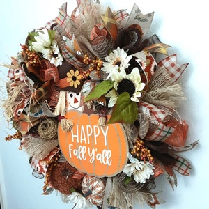 Puede incluir: Una corona de oto&ntilde;o con un letrero en forma de calabaza que dice "HAPPY Fall y'all". La corona presenta girasoles, follaje de oto&ntilde;o y un espantap&aacute;jaros. La paleta de colores incluye naranja, marr&oacute;n y crema.