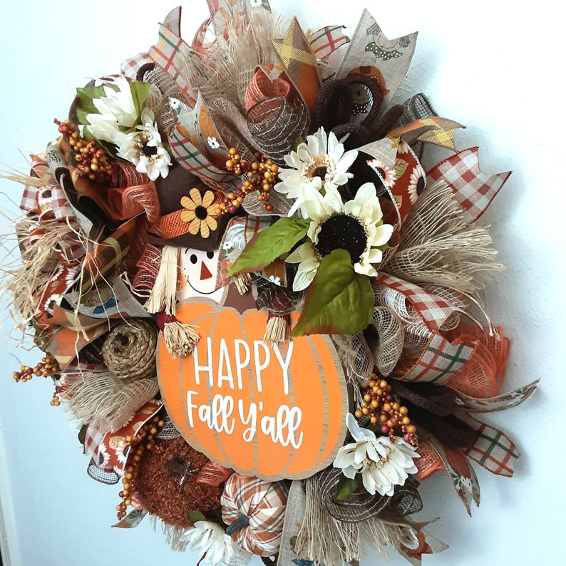 Puede incluir: Una corona de oto&ntilde;o con un letrero en forma de calabaza que dice "HAPPY Fall y'all". La corona presenta girasoles, follaje de oto&ntilde;o y un espantap&aacute;jaros. La paleta de colores incluye naranja, marr&oacute;n y crema.