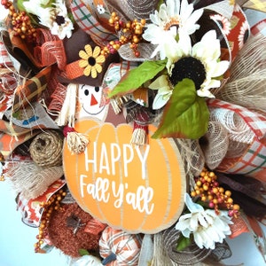Puede incluir: Una corona de oto&ntilde;o con un dise&ntilde;o de espantap&aacute;jaros y calabaza. La corona presenta una gran calabaza naranja con el texto "HAPPY Fall y'all". Est&aacute; decorada con girasoles, hojas de oto&ntilde;o y cintas a cuadros. Los colores son naranja, marr&oacute;n y blanco.