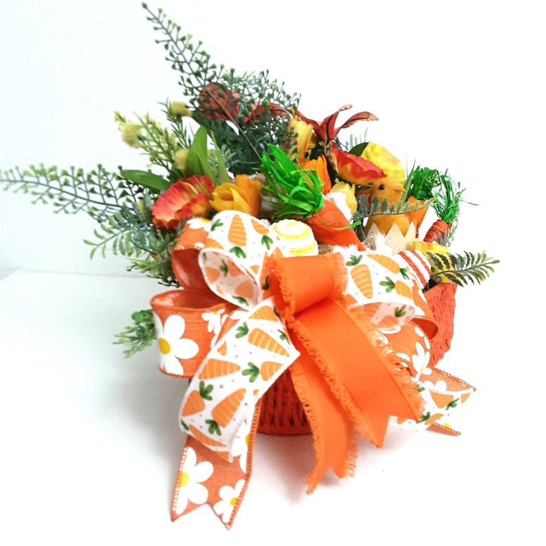 May include: An Easter-themed floral arrangement in a small orange basket. The arrangement includes colourful flowers, greenery, and decorative elements. A large orange bow and a carrot-patterned ribbon add to the festive look. The basket is decorated with white daisies.