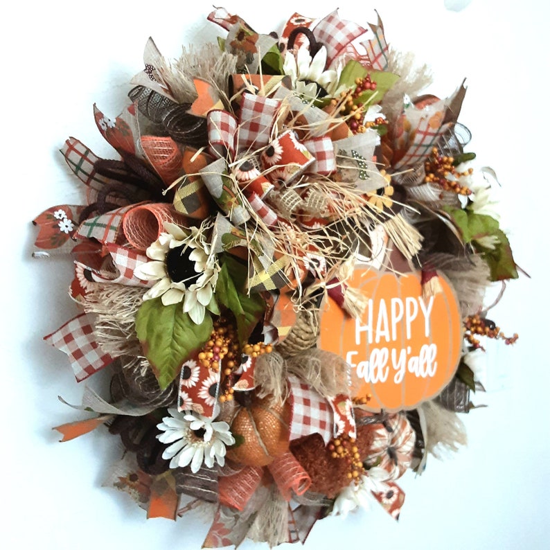 Puede incluir: Una corona de tem&aacute;tica oto&ntilde;al con una variedad de texturas y colores. Presenta girasoles, calabazas y cintas en tonos naranja, marr&oacute;n y crema. Un letrero de madera con forma de calabaza dice "Happy Fall y'all".
