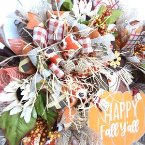 Puede incluir: Una corona con tem&aacute;tica oto&ntilde;al con un dise&ntilde;o de calabaza y espantap&aacute;jaros. La corona presenta cintas naranjas, marrones y blancas, girasoles y el texto "HAPPY Fall y'all". Los elementos decorativos incluyen paja y bayas.