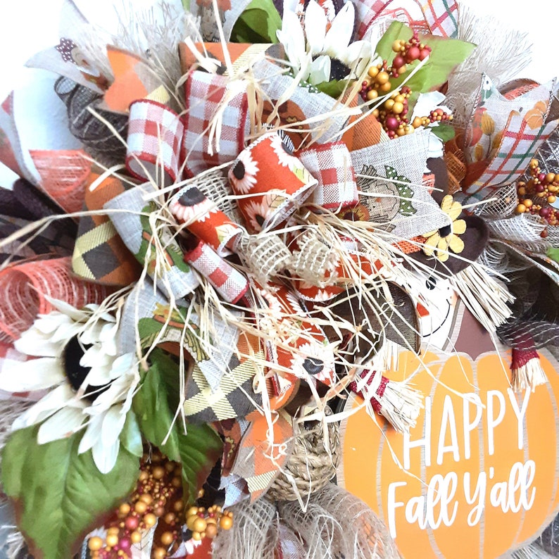 Puede incluir: Una corona con tem&aacute;tica oto&ntilde;al con un dise&ntilde;o de calabaza y espantap&aacute;jaros. La corona presenta cintas naranjas, marrones y blancas, girasoles y el texto "HAPPY Fall y'all". Los elementos decorativos incluyen paja y bayas.