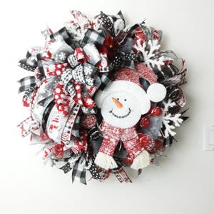Puede incluir: Una corona navide&ntilde;a decorativa con un mu&ntilde;eco de nieve central que lleva un gorro y una bufanda rojos. La corona est&aacute; hecha con capas de cintas a cuadros negros y blancos, rojas y plateadas, decorada con copos de nieve blancos brillantes.