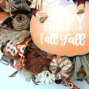 Puede incluir: Una corona de oto&ntilde;o con un gran letrero en forma de calabaza naranja que dice "HAPPY Fall y'all" en letras blancas. La corona incluye calabazas decorativas, girasoles y detalles de arpillera en tonos naranja, marr&oacute;n y blanco.