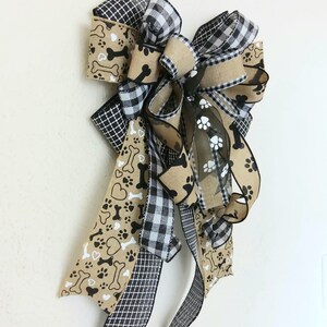 May include: A large, multi-layered bow made with brown burlap, black and white buffalo plaid, and brown ribbon with white paw prints and bone designs. The bow is tied with a black and white checkered ribbon.