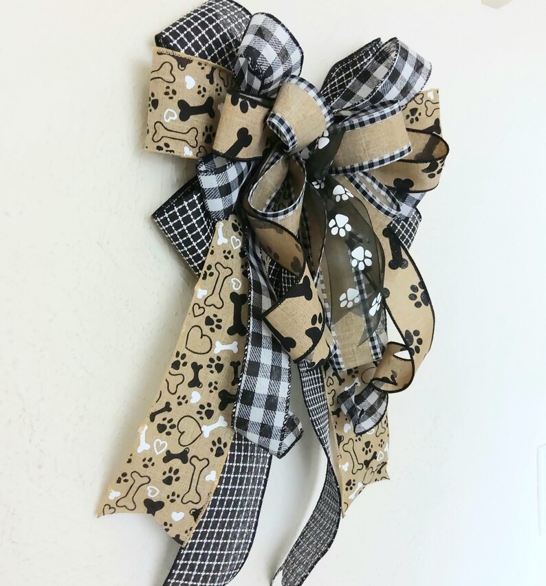 May include: A large, multi-layered bow made with brown burlap, black and white buffalo plaid, and brown ribbon with white paw prints and bone designs. The bow is tied with a black and white checkered ribbon.