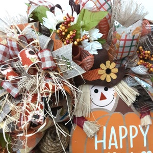 Puede incluir: Una corona de oto&ntilde;o con un dise&ntilde;o de espantap&aacute;jaros. La corona presenta una calabaza con la palabra "HAPPY", un espantap&aacute;jaros con sombrero y varias cintas en patrones naranja, marr&oacute;n y a cuadros. Flores blancas y bayas a&ntilde;aden un toque festivo.