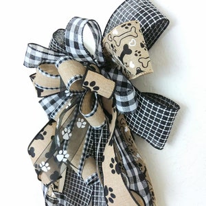 May include: A black and white checkered ribbon, a brown burlap ribbon with black paw prints, and a brown burlap ribbon with white paw prints and bone designs are tied together in a bow.
