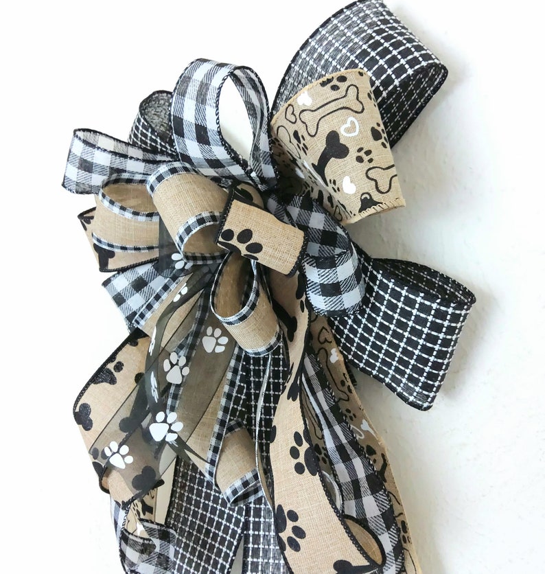 May include: A black and white checkered ribbon, a brown burlap ribbon with black paw prints, and a brown burlap ribbon with white paw prints and bone designs are tied together in a bow.