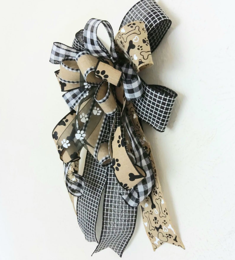 May include: A black and white checkered ribbon, a brown ribbon with white paw prints, and a brown ribbon with white paw prints and bone designs are tied together in a bow.
