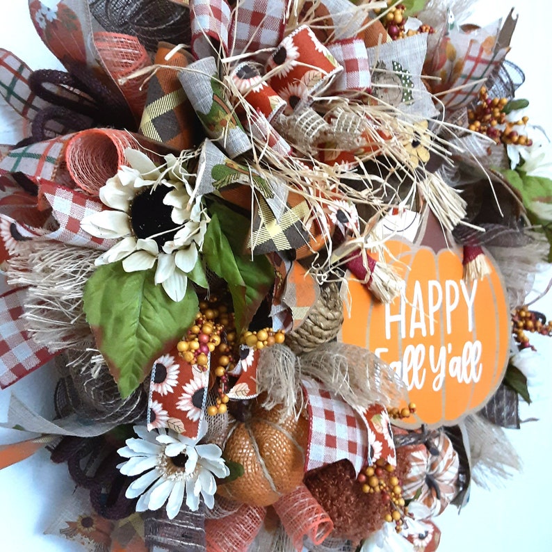 Puede incluir: Una corona de oto&ntilde;o con un letrero en forma de calabaza que dice "Happy Fall y'all". La corona presenta girasoles, calabazas, bayas y varias cintas en naranja, marr&oacute;n y blanco. Est&aacute; decorada con paja y otros elementos de temporada.