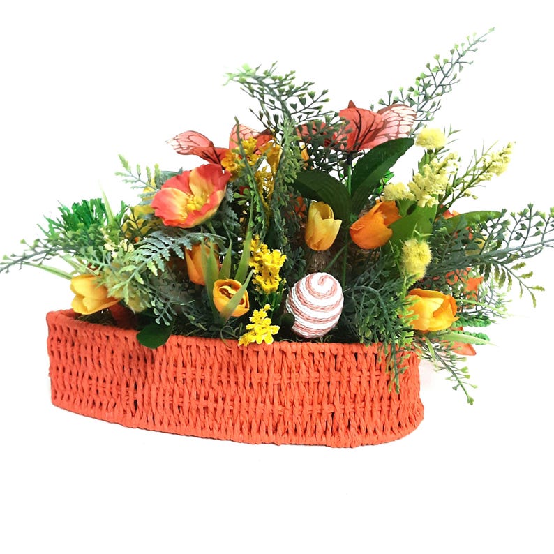 May include: A heart-shaped orange woven basket filled with artificial flowers and foliage. The arrangement features orange tulips, yellow flowers, and Easter eggs. The basket is filled with greenery.