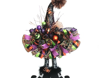 Puede incluir: Un adorno de pared que presenta un sombrero de bruja negro decorado con adornos, cintas y ramitas brillantes en tonos morado, naranja y verde. Debajo del sombrero hay dos piernas con patrones de telaraña y botas negras con hebillas.